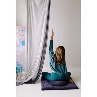 Kobieta siedząca w pozycji medytacyjnej na fioletowej poduszce do medytacji i granatowej macie, podnosi jedną rękę do góry; w tle białe zasłony i kolorowy obraz.